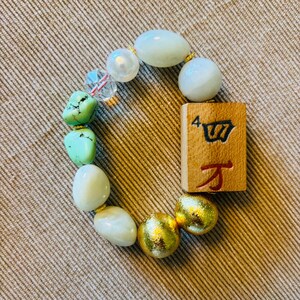 Puede incluir: Una pulsera el&aacute;stica que presenta una ficha de mahjong de madera rectangular con el n&uacute;mero 4 y el car&aacute;cter chino para diez mil, intercalada con cuentas blancas lisas, pepitas de turquesa, dos esferas met&aacute;licas doradas y una cuenta facetada transparente.