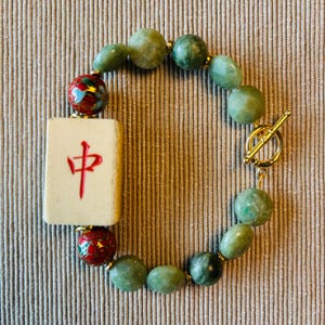 May include: A jade bead bracelet featuring a cream-colored Mahjong tile with a red Chinese character. The bracelet includes green beads, gold accents, and a gold toggle clasp. The beads are approximately 0.5 inches in diameter.