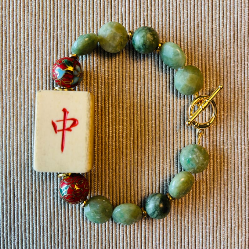 May include: A jade bead bracelet featuring a cream-colored Mahjong tile with a red Chinese character. The bracelet includes green beads, gold accents, and a gold toggle clasp. The beads are approximately 0.5 inches in diameter.