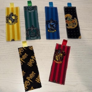 May include: Rectangular fabric bookmarks with various house crests and colors. The bookmarks feature yellow and black stripes, green stripes, blue stripes, black with gold dots, black with gold text, and red stripes. Each bookmark has a ribbon loop.