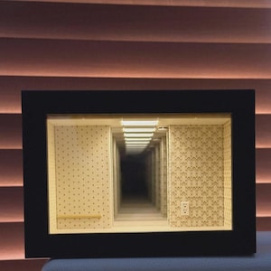 May include: A framed miniature art piece featuring a hallway with repeating walls and ceiling lights, creating an optical illusion of infinite depth. The walls are decorated with patterned wallpaper. The frame is black.