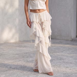 Könnte beinhalten: Cremefarbene Hose mit Rüschen in mehreren Lagen und einem Kordelzug in der Taille. Die Hose wird mit einem passenden Camisole-Top getragen. Der Stoff wirkt leicht und fließend, was auf einen lässigen oder böhmischen Stil hindeutet. Die Hose hat mehrere Rüschen.