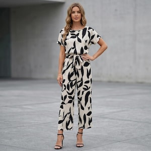 Könnte beinhalten: Cremefarbener Jumpsuit mit schwarzem Blättermuster. Der Jumpsuit hat kurze Ärmel, eine Bindetaille und weite Hosenbeine. Das Model trägt schwarze High-Heel-Sandalen.
