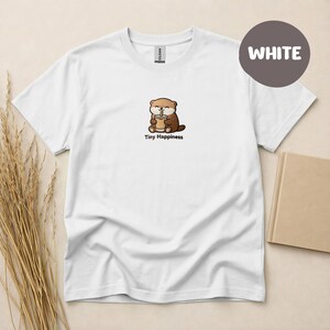Op de afbeelding: Wit T-shirt met een cartoon otter die een drankje vasthoudt en de tekst "Tiny Happiness" eronder. Het shirt heeft een ronde hals en korte mouwen. Een grijze cirkel met het woord "WHITE" staat in de rechterbovenhoek.