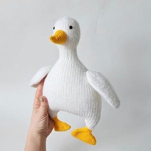 Könnte beinhalten: Ein weißes, gestricktes Entenspielzeug mit gelbem Schnabel und Füßen. Die Ente hat schwarze Augen und kleine Flügel. Das Spielzeug besteht aus einem weichen, strukturierten Material.