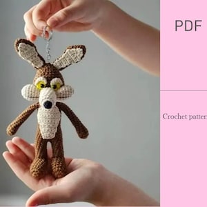 May include: A crocheted keychain in the shape of a cartoon coyote. The coyote is brown with cream-colored accents on the ears, snout, and chest. It has large yellow eyes and is suspended from a silver keyring.