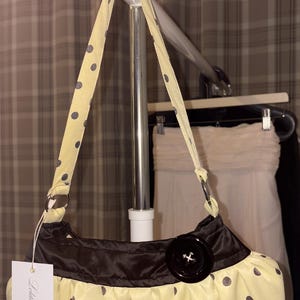 Peut inclure: Un sac à main jaune pâle à pois gris foncé, doté d'une bande supérieure en satin marron foncé, d'un gros bouton noir décoratif et d'une bandoulière assortie à pois. Une étiquette rectangulaire blanche avec le texte Lolita Archive est attachée.