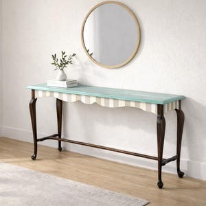 May include: A teal and white striped console table with dark brown legs. The table has a scalloped edge and is topped with a vase of greenery and books. A round mirror hangs on the wall above.