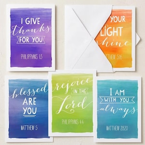 May include: A set of five rectangular greeting cards with watercolor backgrounds in purple, orange, blue, green, and teal. Each card features white text with Bible verses including I GIVE THANKS FOR YOU, LET YOUR LIGHT SHINE, BLESSED ARE YOU, REJOICE IN THE LORD, and I AM WITH YOU ALWAYS.