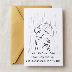 May include: A white greeting card with a black line drawing of two stick figures in the rain. One figure holds an umbrella over the other who is sitting down. The text reads: i can't stop the rain, but i can stand in it with you. A gold envelope.