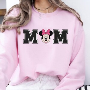 May include: Light pink sweatshirt with the word "MOM" in black block letters, with a Minnie Mouse graphic in the center. The sweatshirt is a solid light pink color. The text "LIGHT PINK" is in a white oval at the bottom.