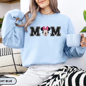 May include: Light blue sweatshirt with the word "MOM" in black block letters, featuring Minnie Mouse's face in the center. The sweatshirt is light blue and has long sleeves. The text "LIGHT BLUE" is at the bottom.