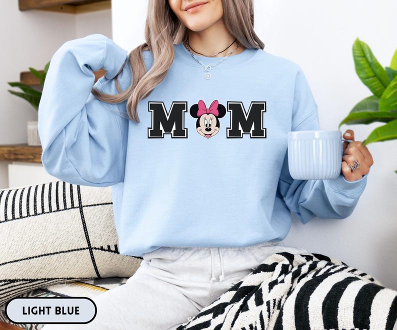 May include: Light blue sweatshirt with the word "MOM" in black block letters, featuring Minnie Mouse's face in the center. The sweatshirt is light blue and has long sleeves. The text "LIGHT BLUE" is at the bottom.