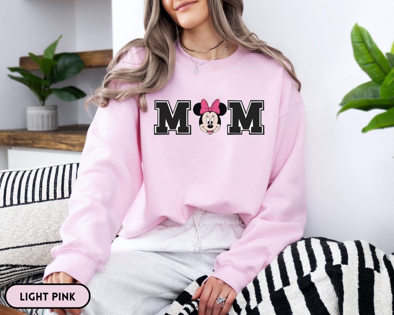 May include: Light pink sweatshirt with the word "MOM" in black block letters, with a Minnie Mouse graphic in the center. The sweatshirt is a solid light pink color. The text "LIGHT PINK" is in a white oval at the bottom.