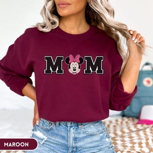 May include: Maroon sweatshirt with the word "MOM" in black block letters with a white outline. The "O" is replaced with a Minnie Mouse graphic wearing a pink bow. The word "MAROON" is printed at the bottom.