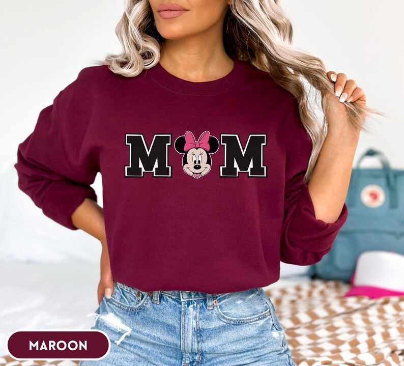 May include: Maroon sweatshirt with the word "MOM" in black block letters with a white outline. The "O" is replaced with a Minnie Mouse graphic wearing a pink bow. The word "MAROON" is printed at the bottom.