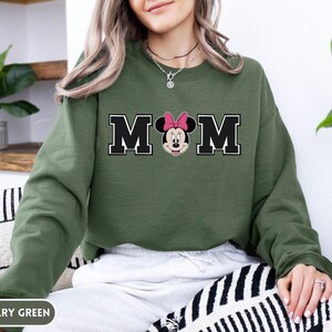 May include: Military green sweatshirt with the word "MOM" in black block letters with a white outline. The "O" is replaced with a Minnie Mouse graphic wearing a pink bow. The sweatshirt is a solid color.