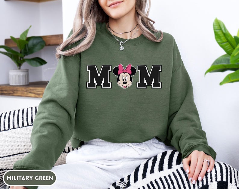 May include: Military green sweatshirt with the word "MOM" in black block letters with a white outline. The "O" is replaced with a Minnie Mouse graphic wearing a pink bow. The sweatshirt is a solid color.