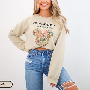 May include: A sand-colored sweatshirt with the word "mama" in black script above the names Nathan, Chole, and Jordan. A floral Minnie Mouse head design with a pink bow is centered on the front.