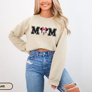 May include: A sand-colored sweatshirt with the word "MOM" in black block letters, featuring a Minnie Mouse graphic. The sweatshirt has long sleeves and a crew neck. The model is wearing blue jeans.