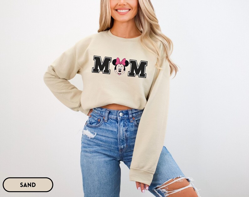 May include: A sand-colored sweatshirt with the word "MOM" in black block letters, featuring a Minnie Mouse graphic. The sweatshirt has long sleeves and a crew neck. The model is wearing blue jeans.