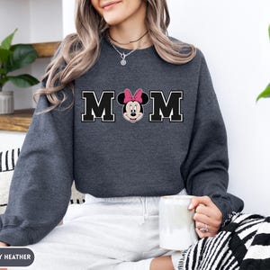 May include: Dark gray heather sweatshirt with the word "MOM" in black block letters with a white outline. The "O" is replaced with a Minnie Mouse graphic wearing a pink bow. The sweatshirt is a long-sleeved crew neck.