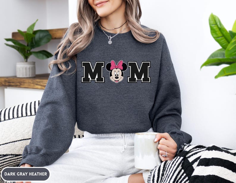 May include: Dark gray heather sweatshirt with the word "MOM" in black block letters with a white outline. The "O" is replaced with a Minnie Mouse graphic wearing a pink bow. The sweatshirt is a long-sleeved crew neck.