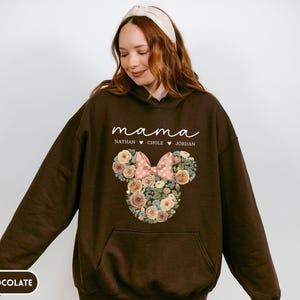 May include: Dark chocolate brown hoodie with a floral Minnie Mouse design. The design includes a pink bow and the word "mama" in white script, along with three names. The hoodie has a front pocket and a hood.