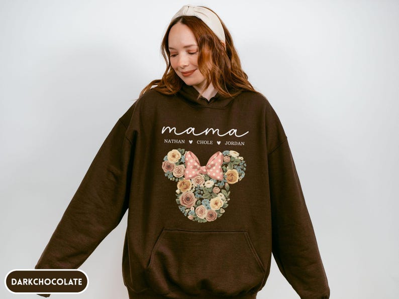 May include: Dark chocolate brown hoodie with a floral Minnie Mouse design. The design includes a pink bow and the word "mama" in white script, along with three names. The hoodie has a front pocket and a hood.