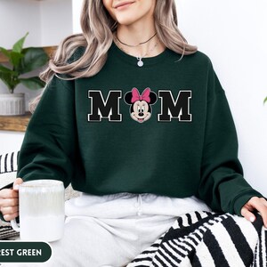 May include: Forest green sweatshirt with the word "MOM" in black block letters with a white outline. The "O" is replaced with a Minnie Mouse graphic wearing a pink bow. The sweatshirt is a crew neck style.