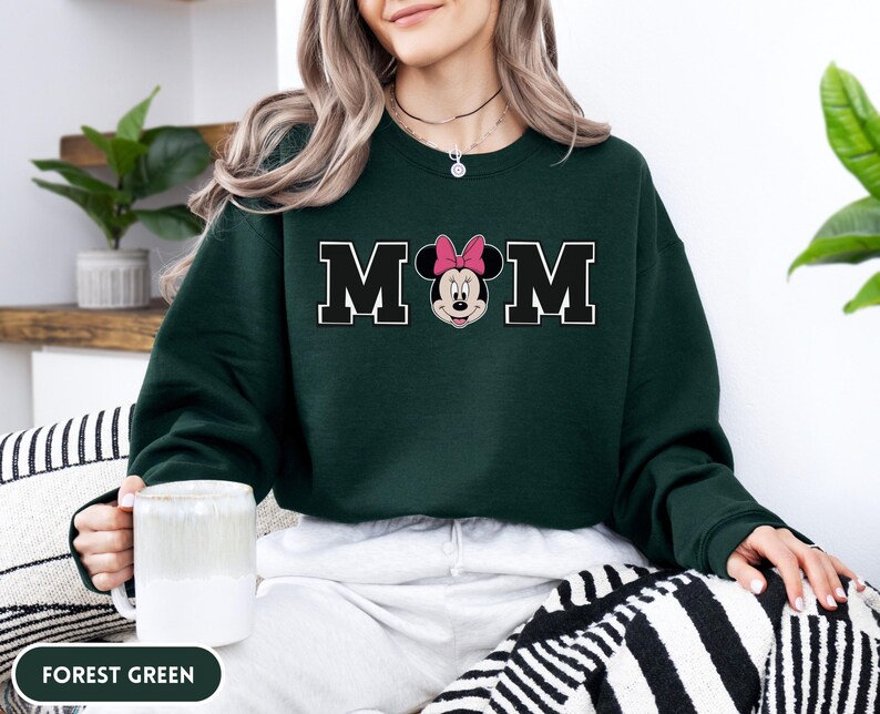 May include: Forest green sweatshirt with the word "MOM" in black block letters with a white outline. The "O" is replaced with a Minnie Mouse graphic wearing a pink bow. The sweatshirt is a crew neck style.