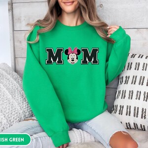 May include: Irish green sweatshirt with the word "MOM" in black block letters with a white outline. The "O" is replaced with a Minnie Mouse graphic wearing a pink bow. The sweatshirt is a solid color.