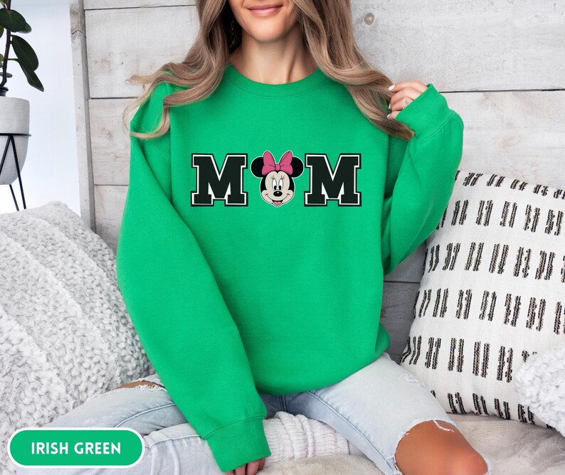 May include: Irish green sweatshirt with the word "MOM" in black block letters with a white outline. The "O" is replaced with a Minnie Mouse graphic wearing a pink bow. The sweatshirt is a solid color.