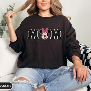May include: Dark chocolate-colored sweatshirt with the word "MOM" in white block letters. The "O" is replaced with a Minnie Mouse graphic wearing a pink bow. The sweatshirt is a long-sleeved crew neck.