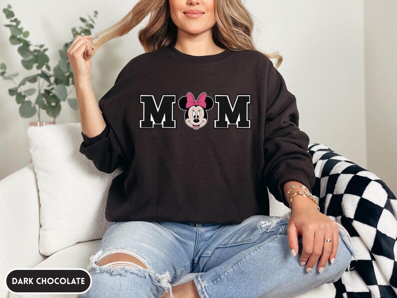 May include: Dark chocolate-colored sweatshirt with the word "MOM" in white block letters. The "O" is replaced with a Minnie Mouse graphic wearing a pink bow. The sweatshirt is a long-sleeved crew neck.