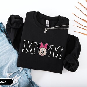 May include: Black sweatshirt with the word "MOM" in white block letters, featuring a Minnie Mouse graphic with a pink bow. A silver necklace is draped across the sweatshirt. The word "BLACK" is in a black rectangle at the bottom.