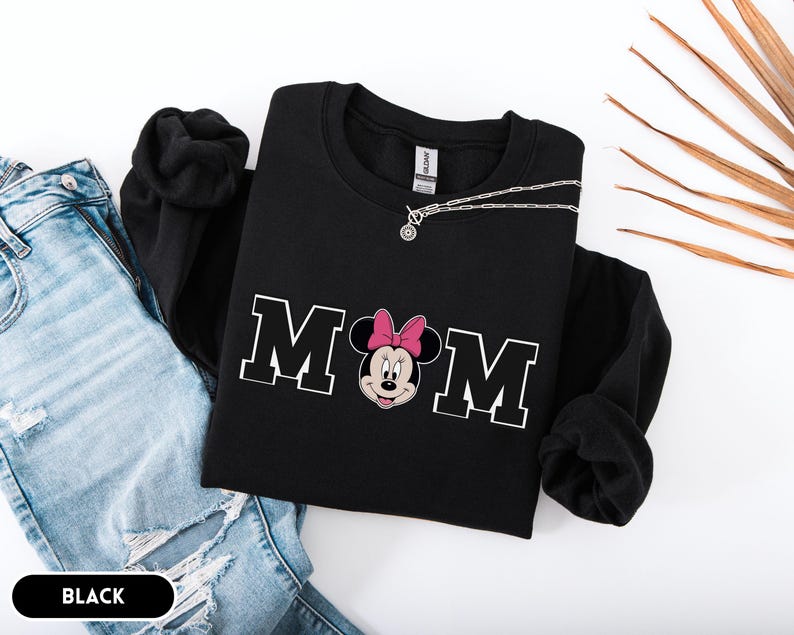 May include: Black sweatshirt with the word "MOM" in white block letters, featuring a Minnie Mouse graphic with a pink bow. A silver necklace is draped across the sweatshirt. The word "BLACK" is in a black rectangle at the bottom.