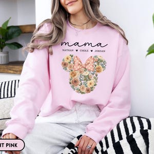 May include: Light pink sweatshirt with the word "mama" in cursive script above a floral Mickey Mouse head design. The design includes a bow and the names Nathan, Chole, and Jordan. The sweatshirt is a light pink color.