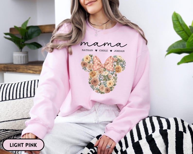 May include: Light pink sweatshirt with the word "mama" in cursive script above a floral Mickey Mouse head design. The design includes a bow and the names Nathan, Chole, and Jordan. The sweatshirt is a light pink color.