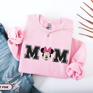 May include: Light pink sweatshirt with the word "MOM" in black block letters with a Minnie Mouse graphic. The sweatshirt is paired with blue jeans. A delicate necklace is draped on the sweatshirt.