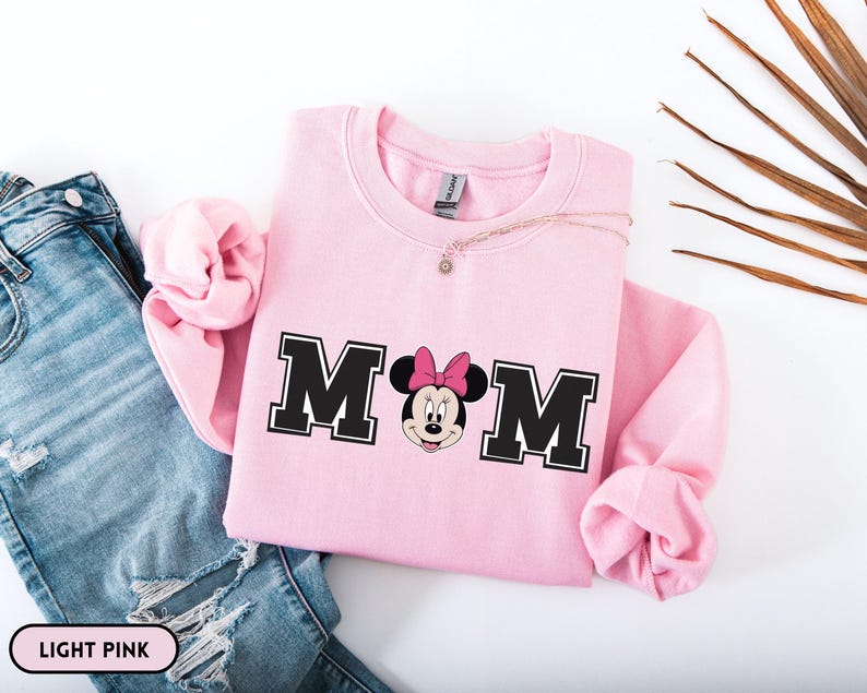 May include: Light pink sweatshirt with the word "MOM" in black block letters with a Minnie Mouse graphic. The sweatshirt is paired with blue jeans. A delicate necklace is draped on the sweatshirt.