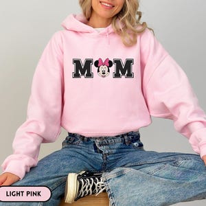 May include: Light pink hooded sweatshirt with the word "MOM" in black block letters, featuring Minnie Mouse in the center. The sweatshirt is a solid light pink color and is paired with blue jeans.