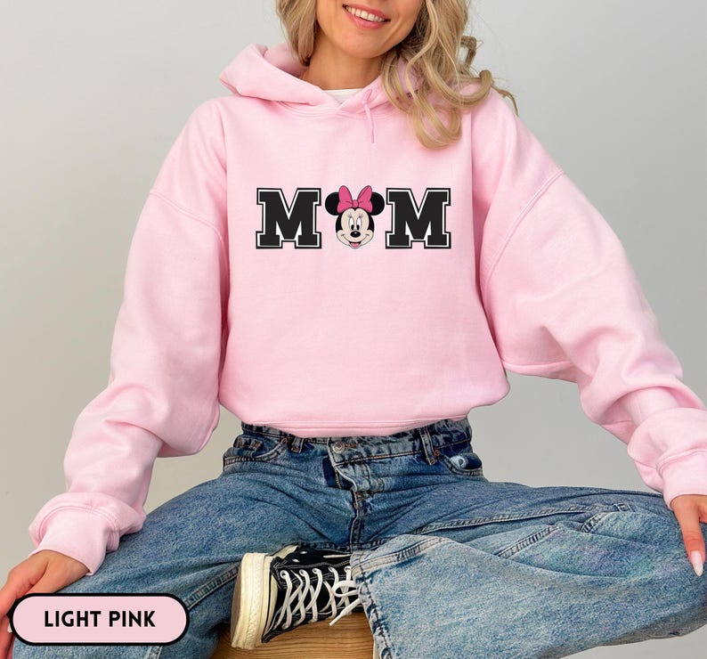 May include: Light pink hooded sweatshirt with the word "MOM" in black block letters, featuring Minnie Mouse in the center. The sweatshirt is a solid light pink color and is paired with blue jeans.