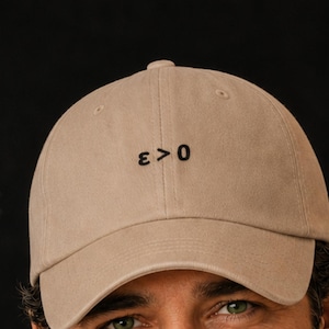 May include: A tan baseball cap with the mathematical symbol "ε > 0" embroidered in black on the front. The cap is made of a soft fabric and has a curved brim. The background includes a chalkboard with mathematical equations.