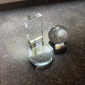 Puede incluir: Una colección de tres piezas de exhibición de acrílico transparente que consta de un bloque rectangular, un disco circular y una esfera lisa. Estos objetos transparentes están dispuestos juntos, mostrando sus superficies pulidas y formas.