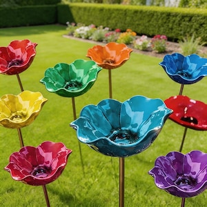 Puede incluir: Un conjunto de adornos de jardín con forma de flor de cerámica colorida. Las flores son de color rojo, naranja, amarillo, verde, azul, turquesa y morado, cada una con un centro oscuro. Están montadas sobre finos tallos metálicos marrones.