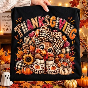 Peut inclure: Un t-shirt noir avec le mot THANKSGIVING en lettres colorées et à motifs au-dessus d'une dinde de dessin animé portant un grand nœud léopard et à carreaux, entourée de tournesols, de citrouilles et de feuilles d'automne.