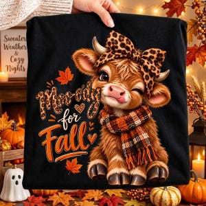 Peut inclure: Un t-shirt noir avec une illustration de vache highland brune portant un nœud à motif léopard et une écharpe à carreaux. Le texte indique Moo-ody for Fall, accompagné de feuilles d'érable orange et de petits cœurs sur le devant.