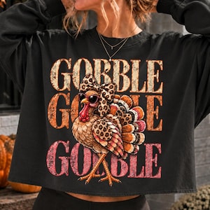 Peut inclure: Un sweat-shirt noir à manches longues avec un motif de dinde portant des lunettes de soleil et un nœud à imprimé léopard. La dinde a des plumes léopard et le texte GOBBLE GOBBLE GOBBLE est imprimé en grandes lettres multicolores usées.