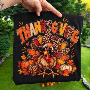 Peut inclure: Un t-shirt noir avec un motif de dinde portant des lunettes de soleil et un nœud à imprimé léopard. Le mot THANKSGIVING est imprimé au-dessus dans une police usée multicolore. Le design inclut des citrouilles et des feuilles d'automne.
